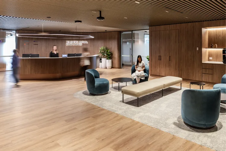 A photo of the lobby of the TMBL head office. It is a modern lobby with wooden ceiling and floors, blue chairs, a long beige bench, and green plants. A reception desk is in the background.