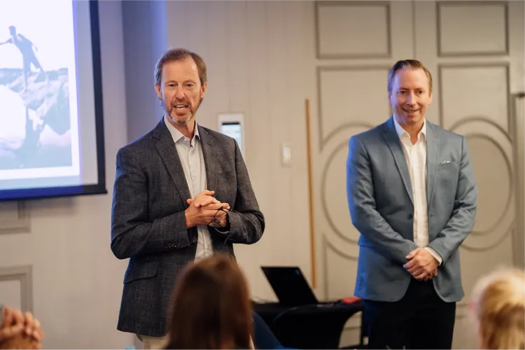 Andrew Kearnan, Chair of the TMBL Board, and Anthony Hughes, TMBL's CEO, stand in a conference room. Andrew is speaking, gesturing with his hands, while Andrew watches approvingly.