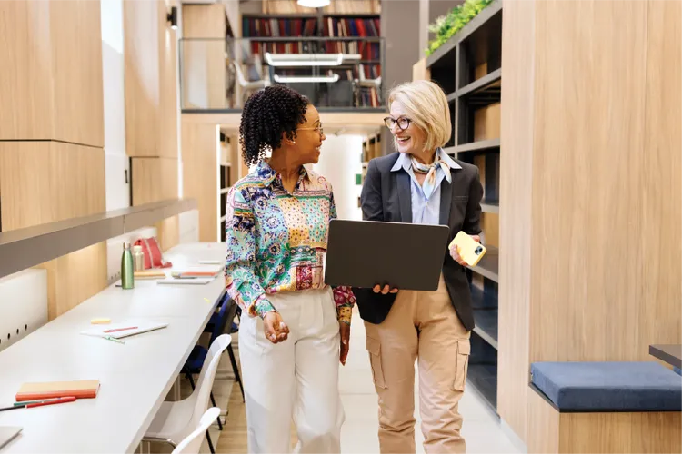 Two professionals walk through a modern, cozy office space. They are engaged in conversation, carrying a laptop and notebooks, conveying a collaborative atmosphere.
