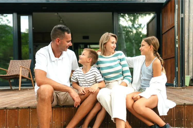 A family of four sits on a wooden deck, smiling and talking in front of their modern house with large glass doors.